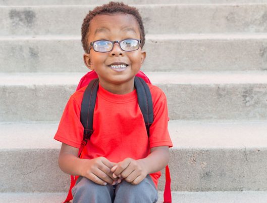 Little kid wearing glasses and a backpack sitting on some stairs.