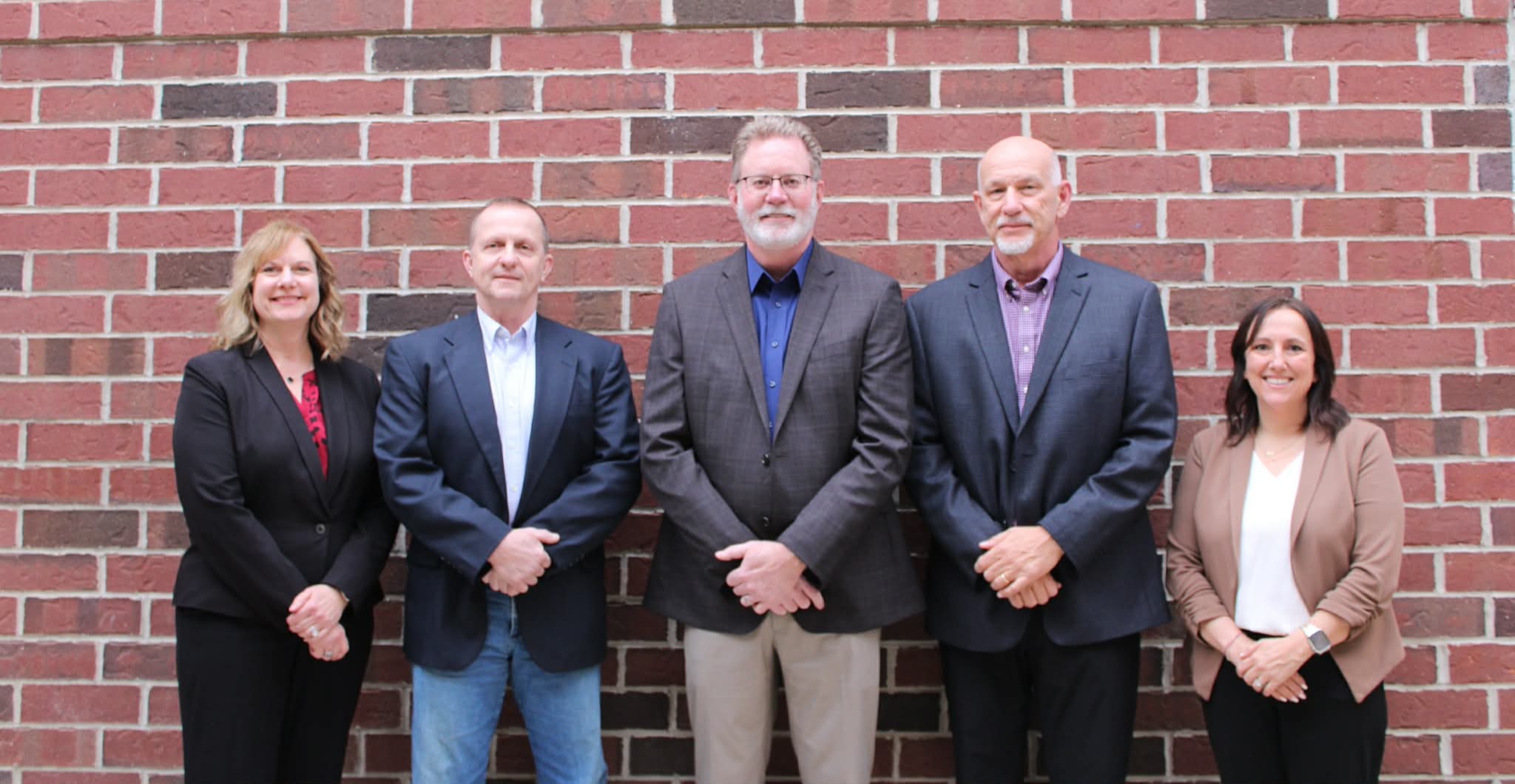Five people in formal attire stand in front of a brick wall. Three men and two women, all smiling.