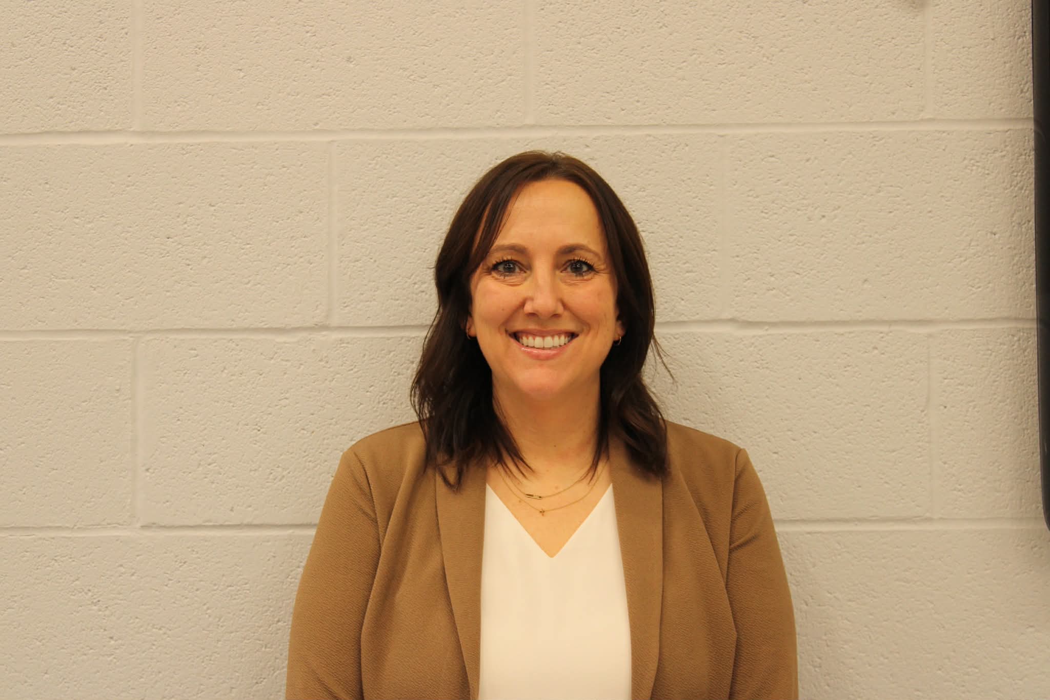 Woman in a white shirt and brown jacket smiles at the camera against a white wall.