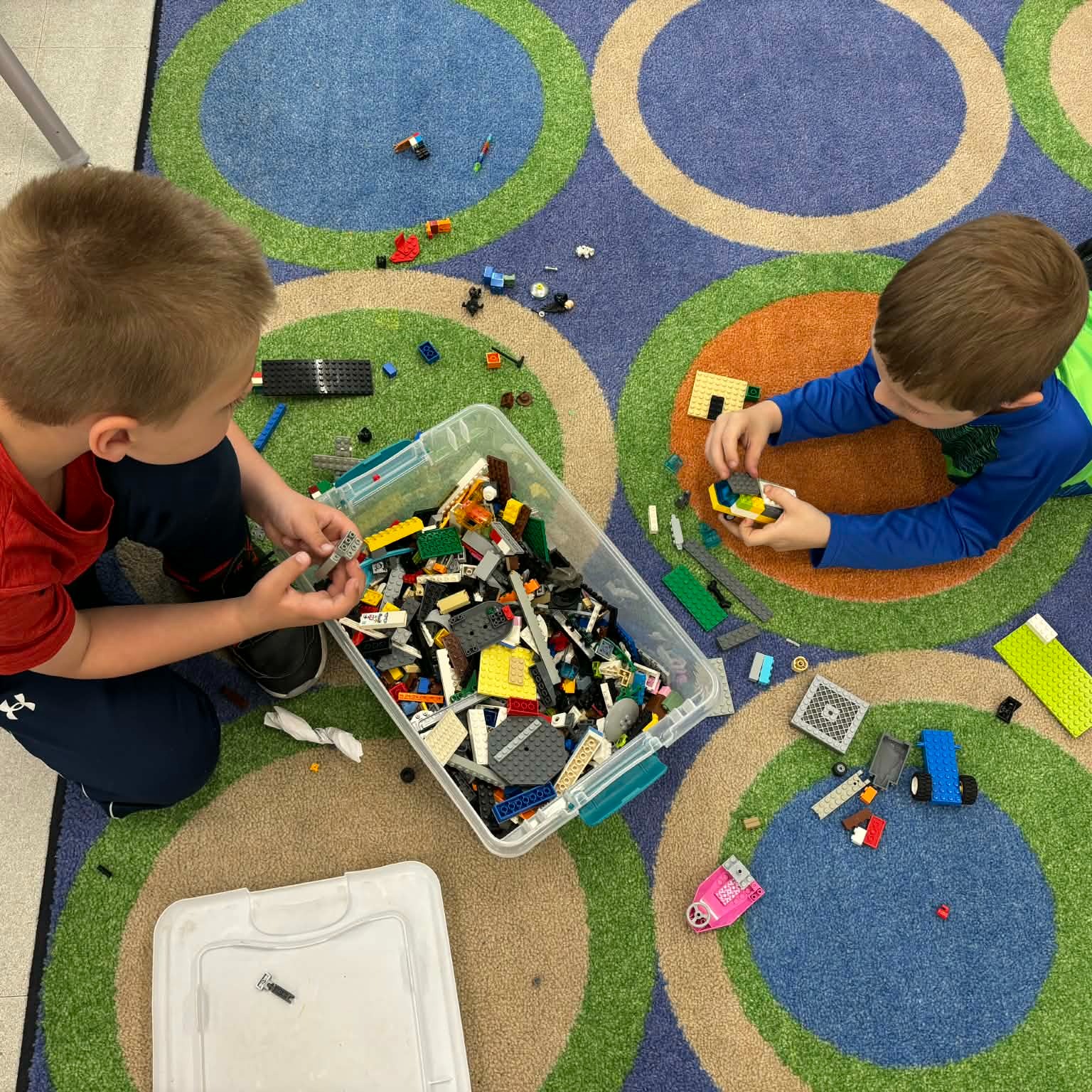 Two children are on a patterned rug, one building with Legos from a box and the other holding a Lego.