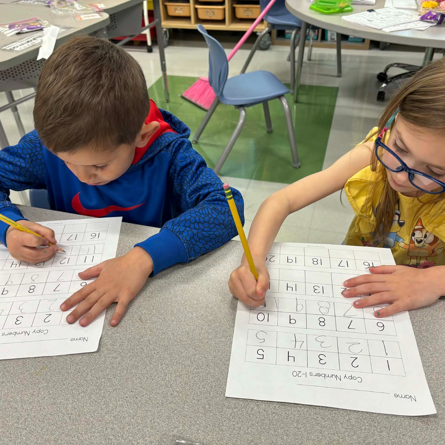 Two children focus on their worksheets, each holding a pencil. One child wears a blue jacket, and the other wears glasses. Behind them are desks and chairs.