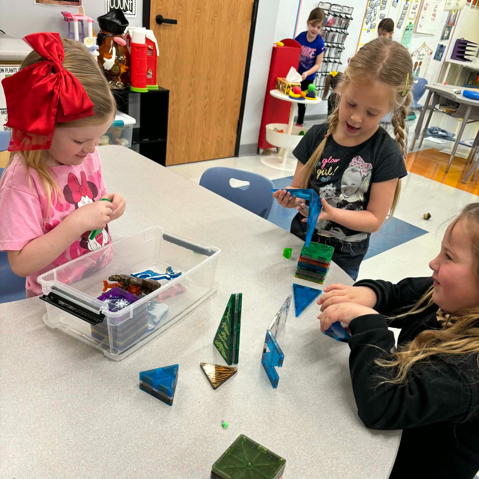 Three girls in a classroom setting, working with triangular-shaped objects on a table.