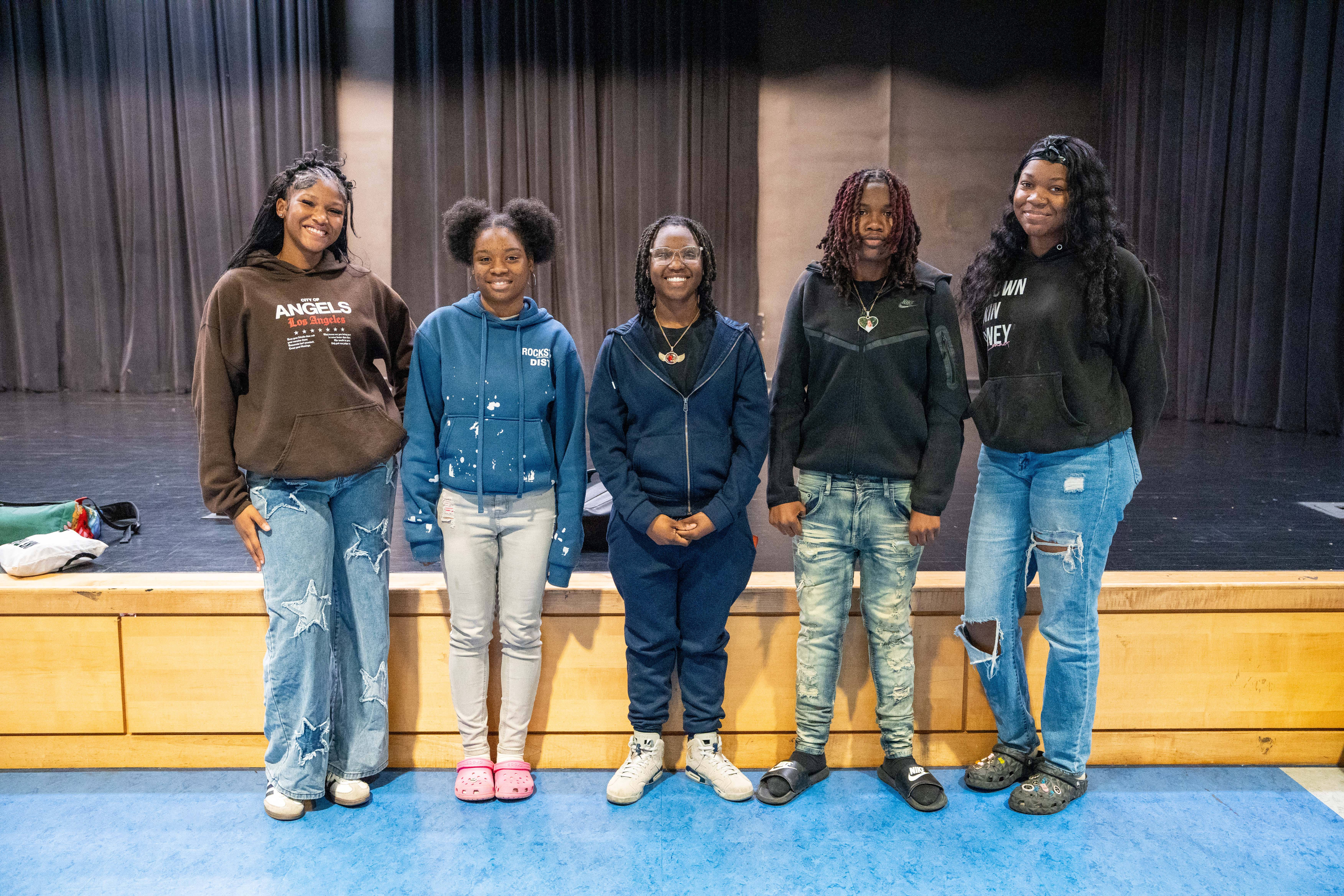 Five women stand together in front of a stage. They wear jeans and jackets. The floor is blue.