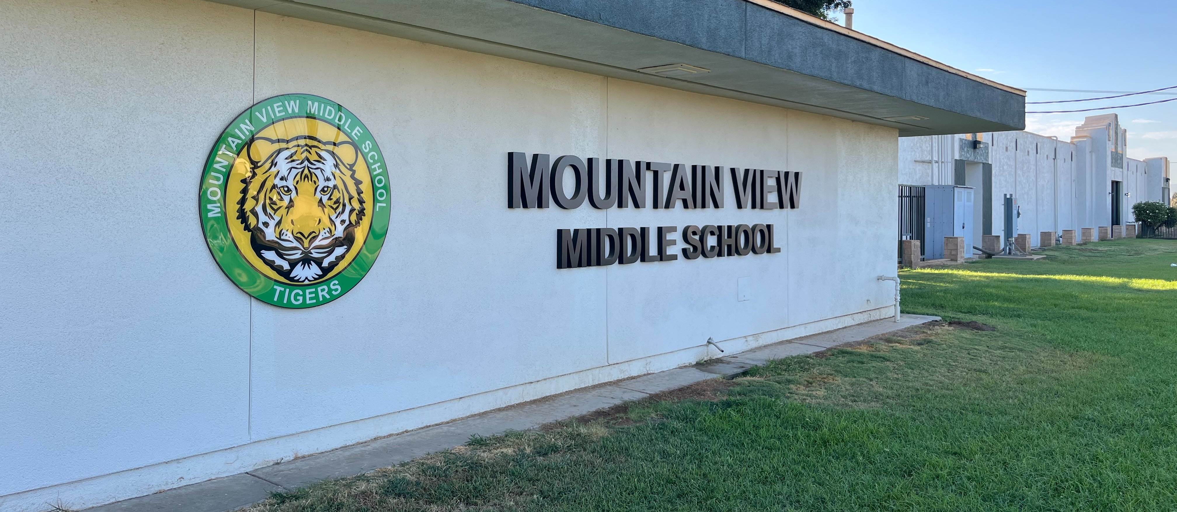 "Mountain View Middle School exterior sign featuring the school's logo with a tiger head and the words 'Mountain View Middle School' in large black letters on a white building wall. The school's emblem includes a circular green and yellow border with the words 'Mountain View Middle School Tigers' surrounding the tiger head.