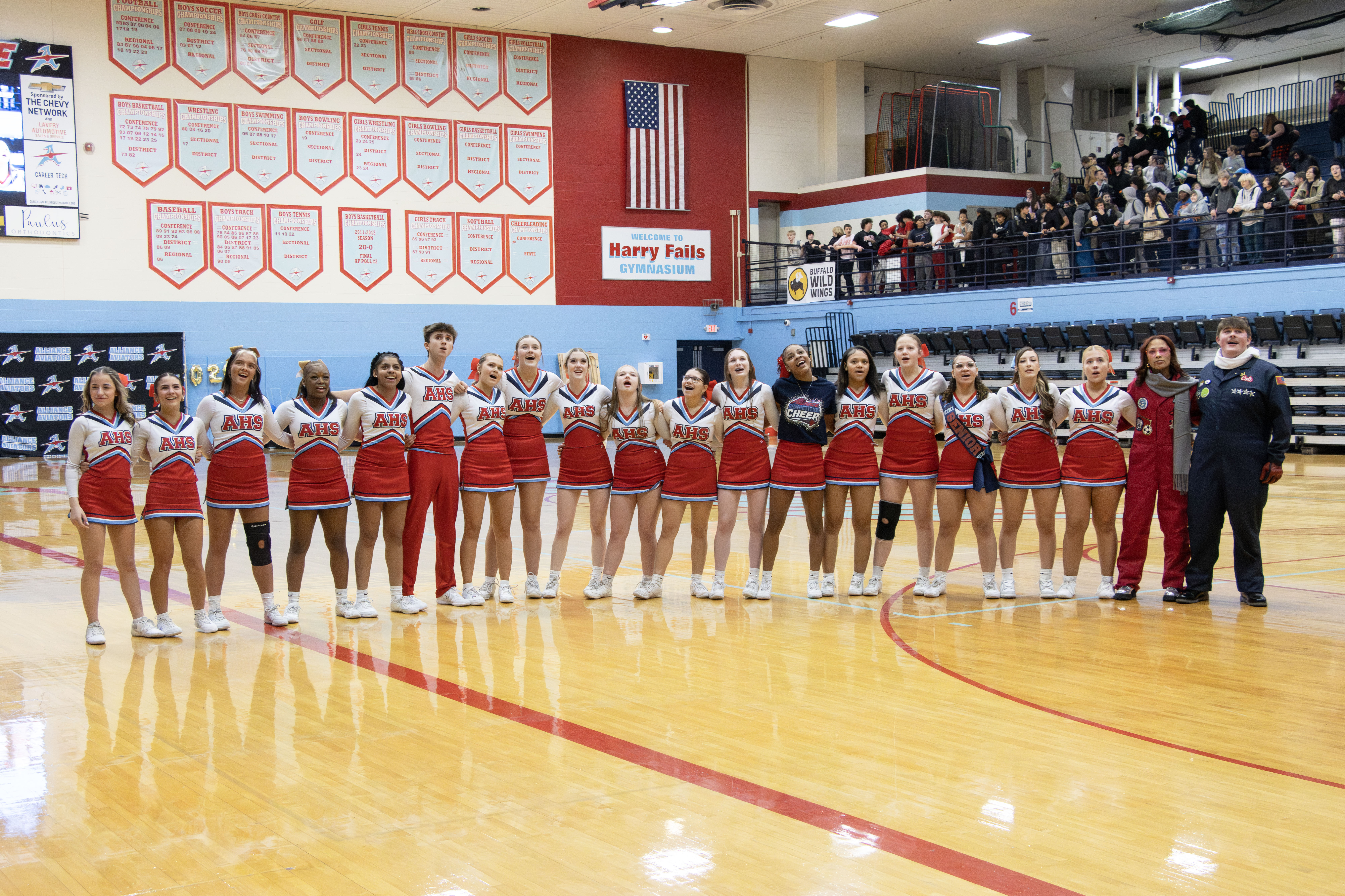 Alliance High School cheerleaders and coaches stand together on the gym floor during a school event, with students watching from the bleachers in the background.