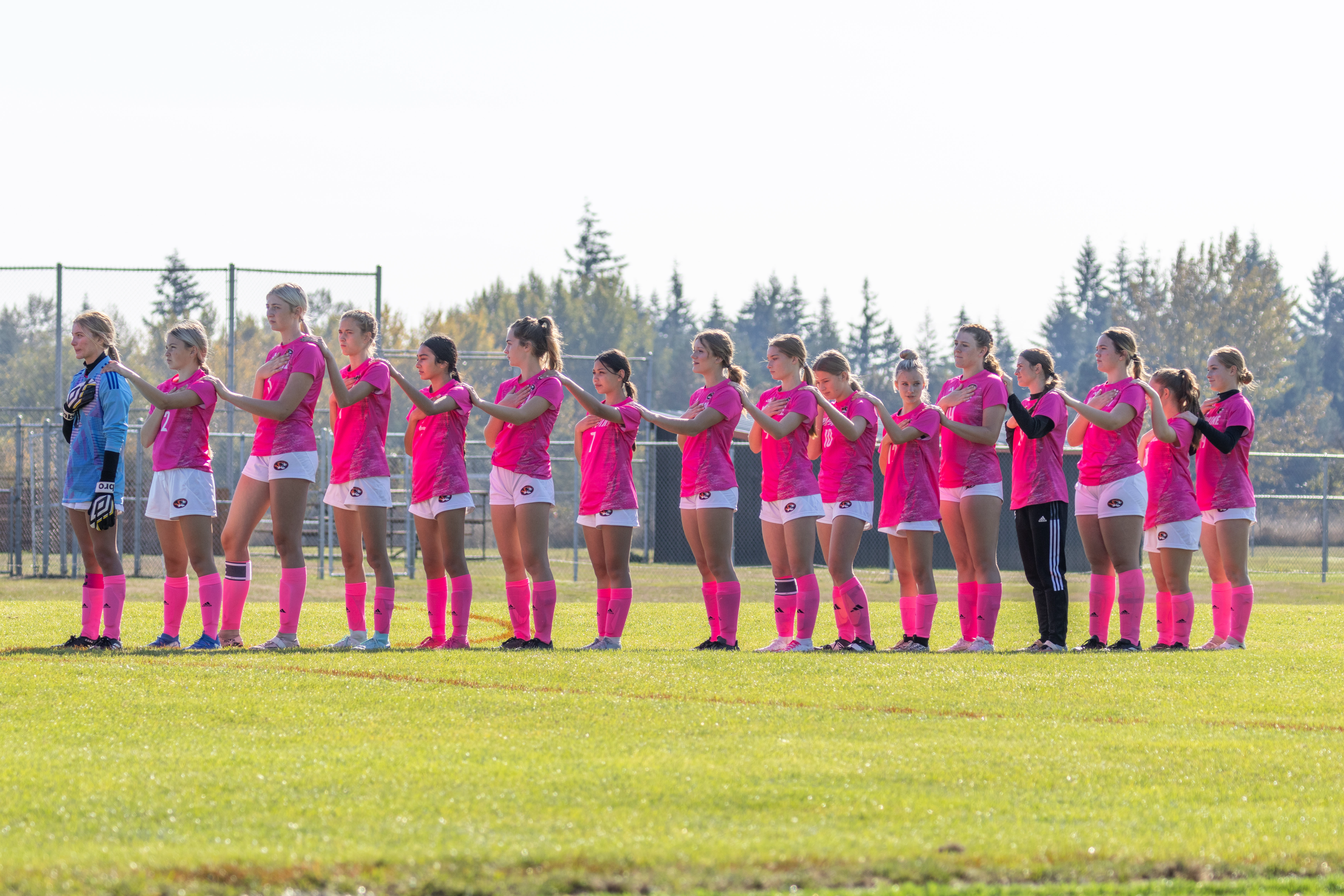 The Napavine High School Girls Soccer team stands lined up together for the National Anthem just before a home match during the day. The goalie is wearing her blue keepers jersey, while the rest of the team is wearing a special "shock pink" jersey with white numers, white shorts and "shock pink" socks, all by Adidas, for Breast Cancer Awareness month.