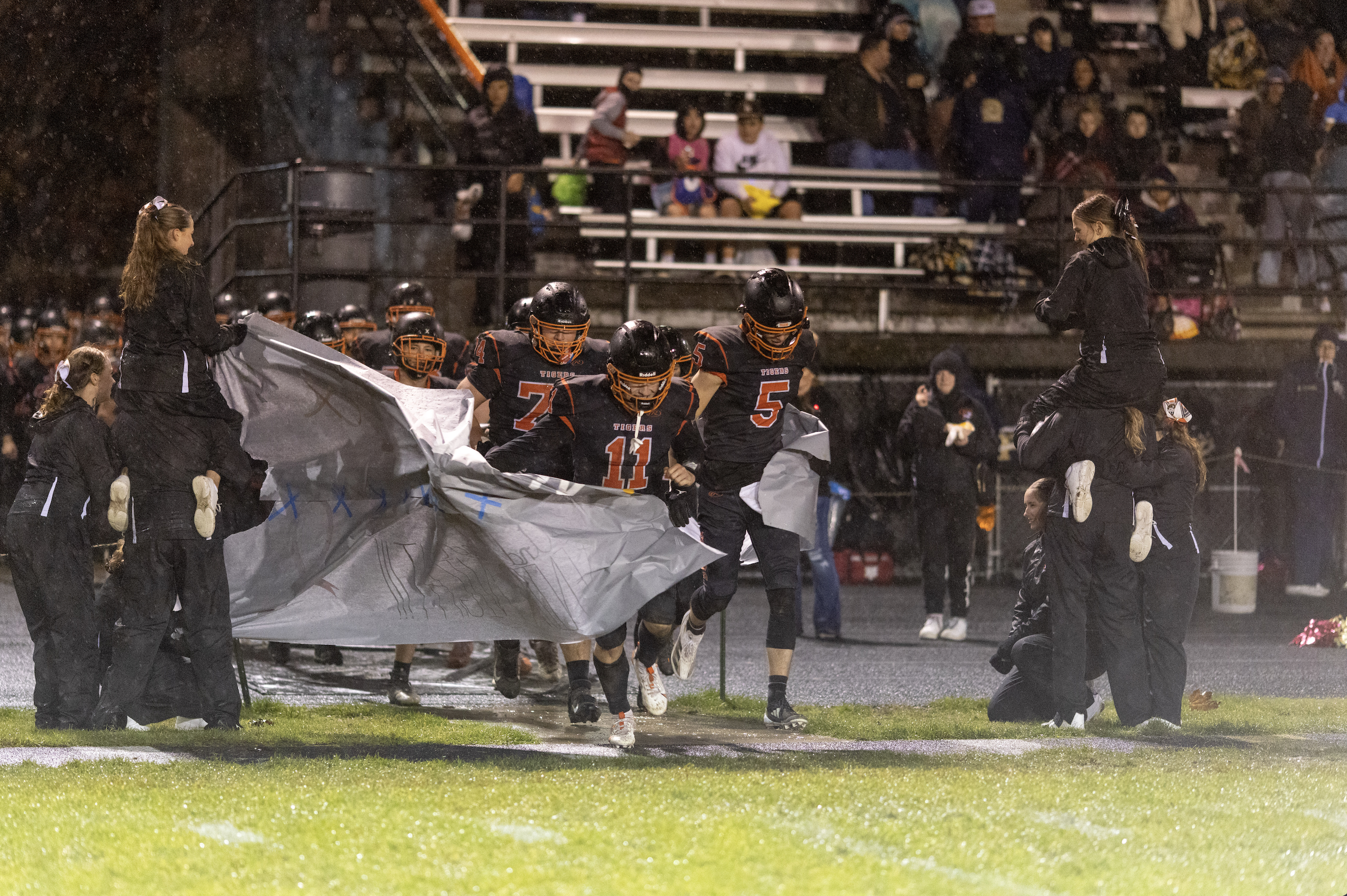 The Napavine High School Football team runs on to the field at the end of halftime on Senior Night. The players are running through large paper sign supporting the team, made and held by the cheerleading squad. The team is wearing their black home uniform with orange lettering and white outline, black helmets and orange facemasks.