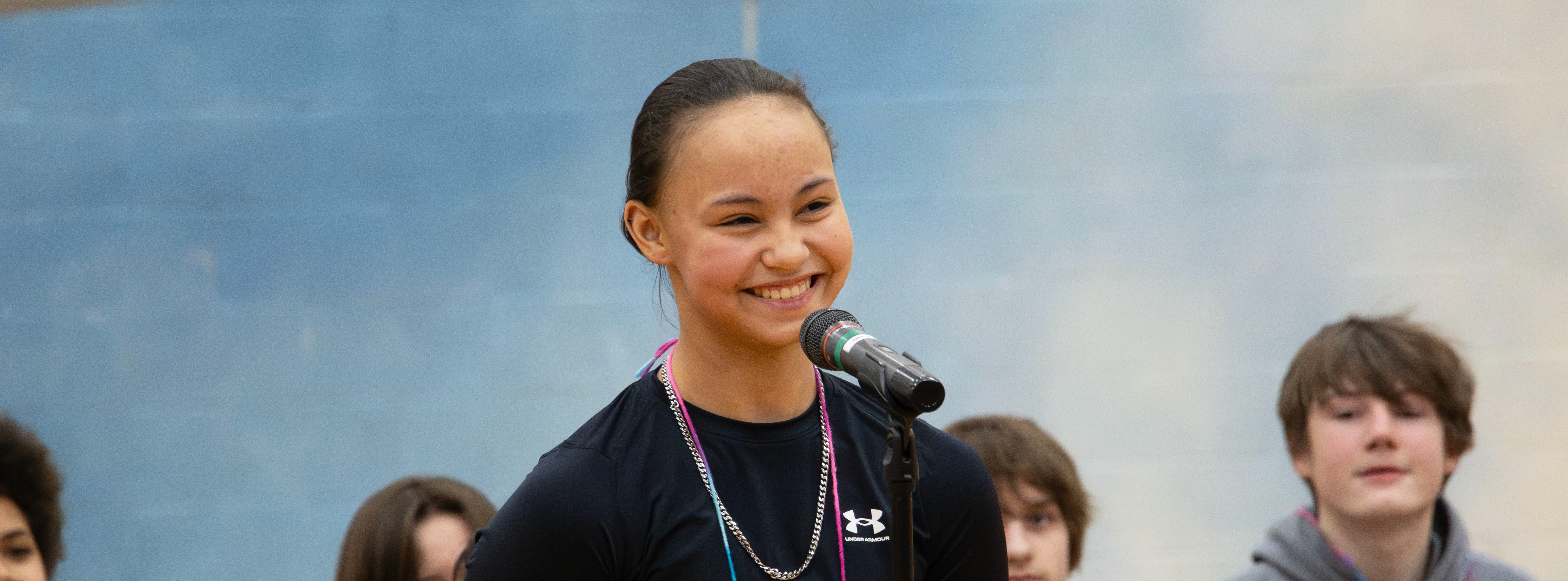 A smiling student stands at a microphone during a school event while other students sit behind her.