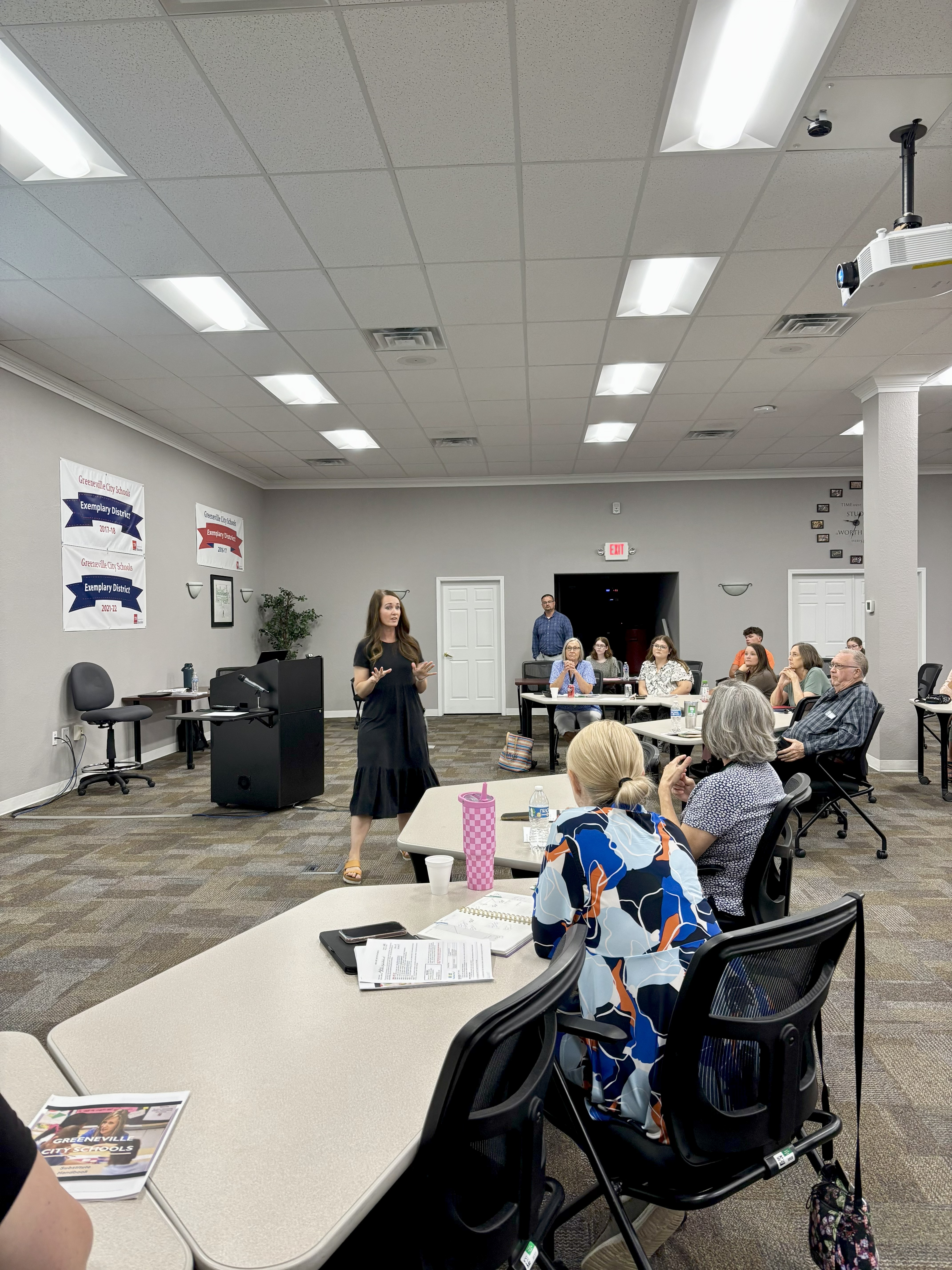 A group of people seated around U-shaped tables in a conference room, attentively listening to a woman speaking at the front. The room is lit with fluorescent lights and decorated with motivational posters reading 'Work Hard' and 'Stay Humble.' A ceiling-mounted projector is visible.