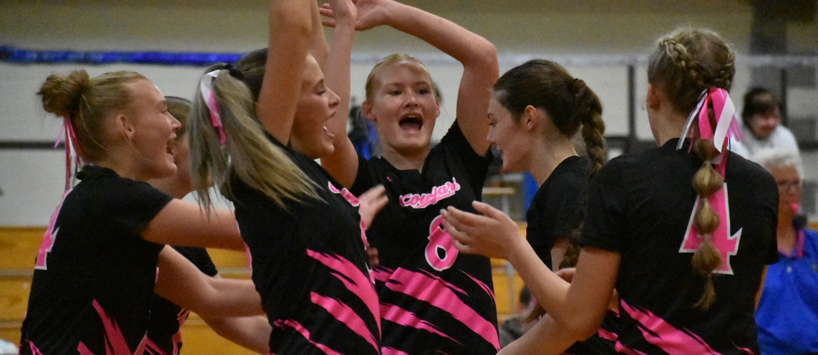 Curlew volleyball players celebrate together on the court while wearing black and pink uniforms in recognition of Breast Cancer Awareness Month.
