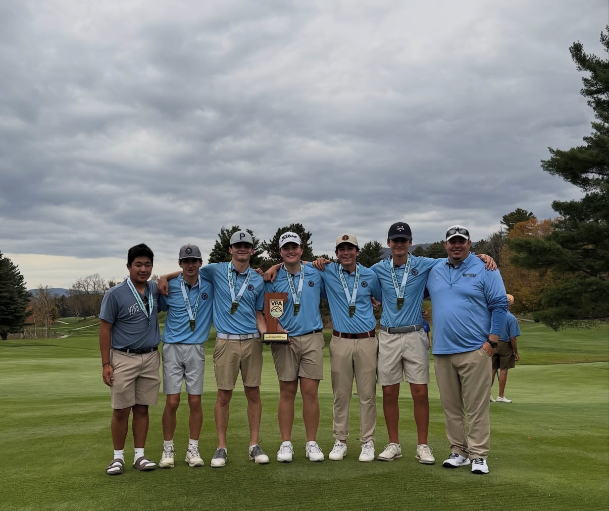Six men stand on a golf course with a trophy, holding it between them. All wear blue shirts and khaki shorts.