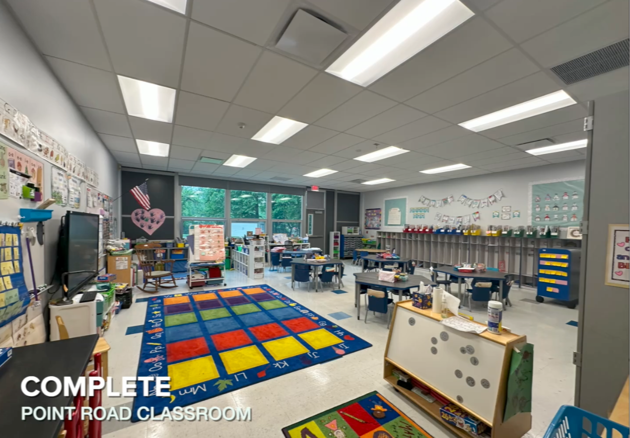Overview of a 1st grade classroom with tables, chairs, a rug, and cabinets adjacent to a bank of windows.