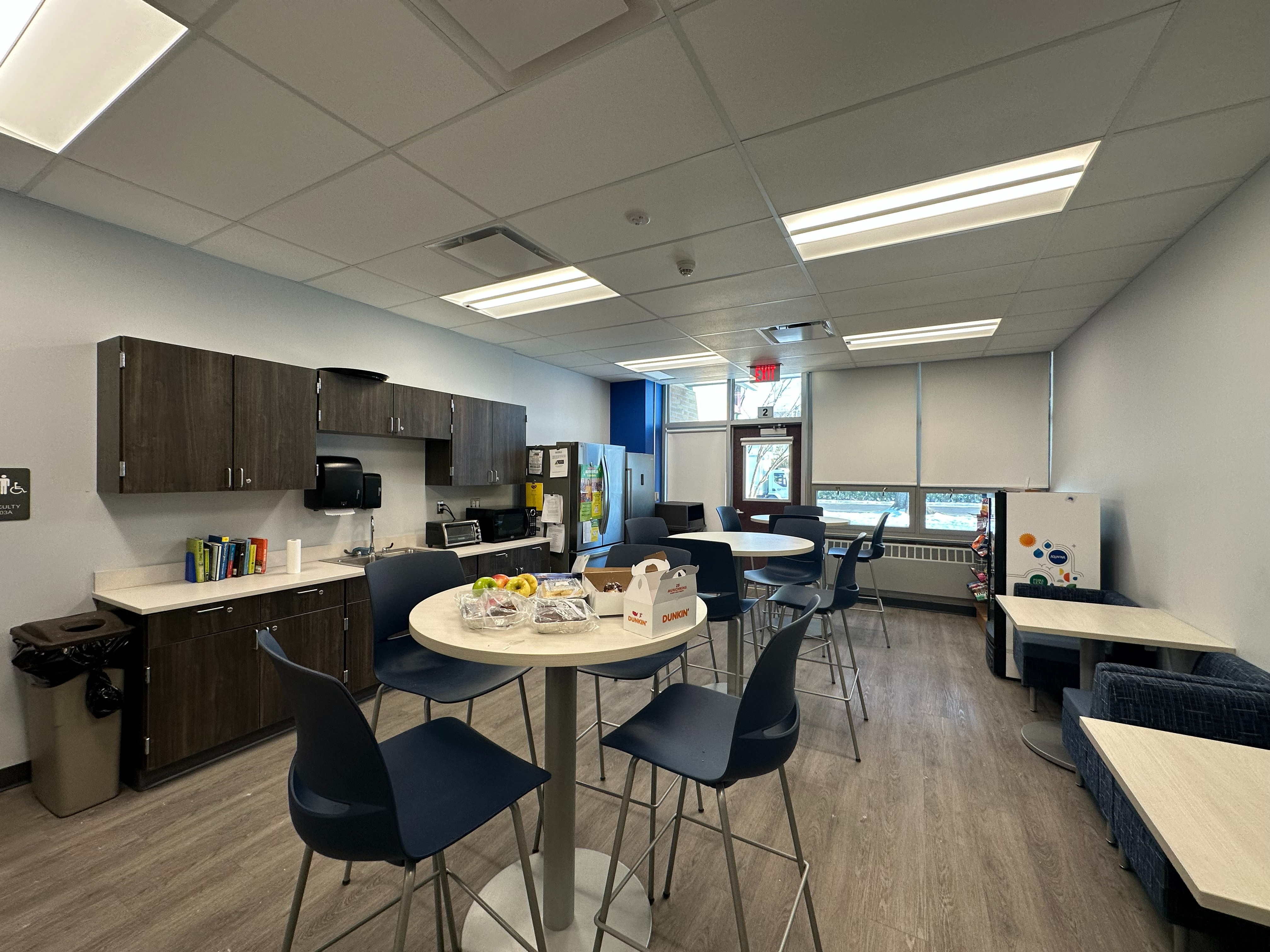 overview of faculty room with high top tables, smaller tables and chairs, as well as a refrigerator and counters with upper and lower cabinets and a sink.