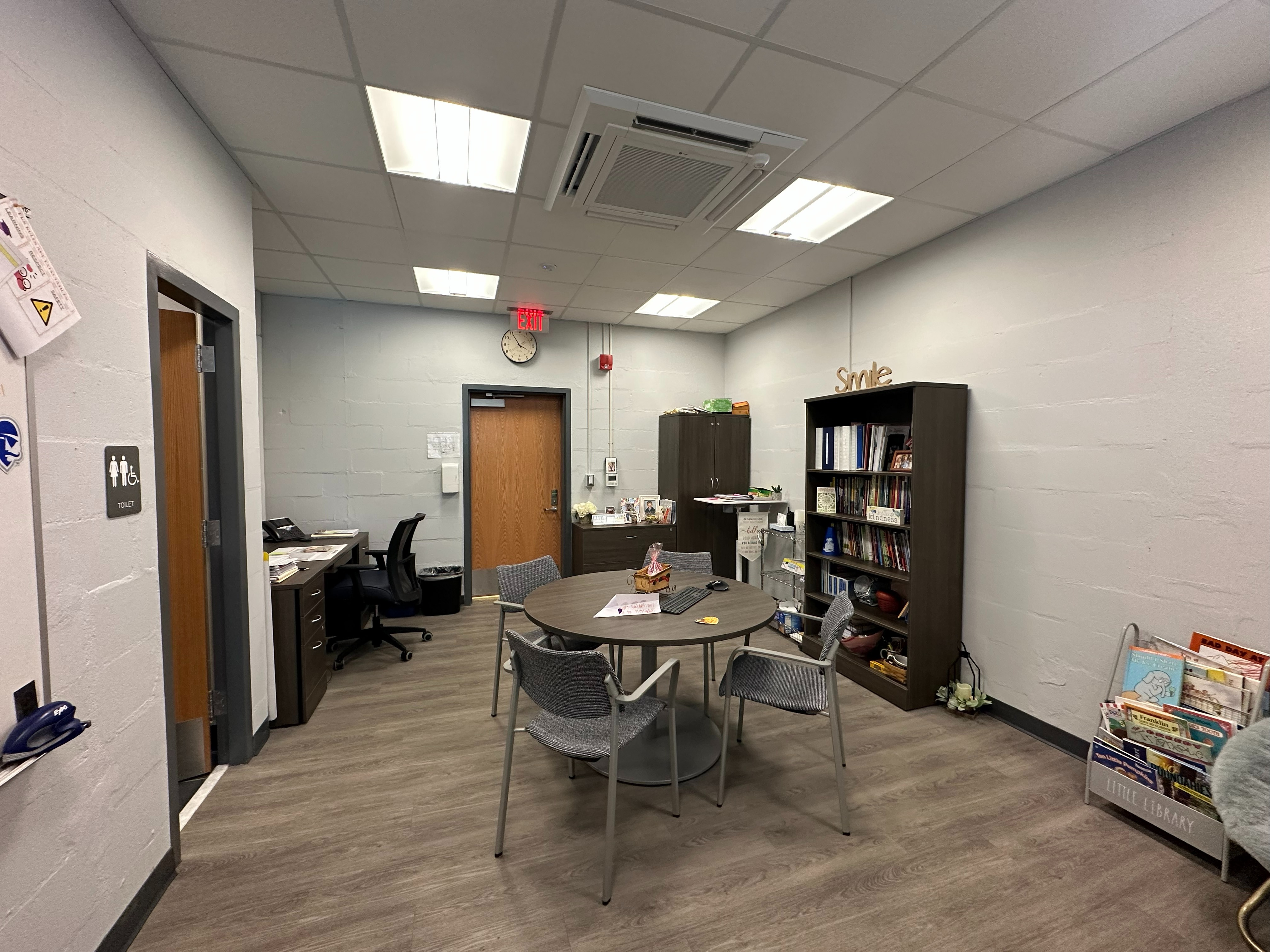 overview of principal's office of small table with 4 chairs, a bookkcase, a door, a desk, and a bathroom door.