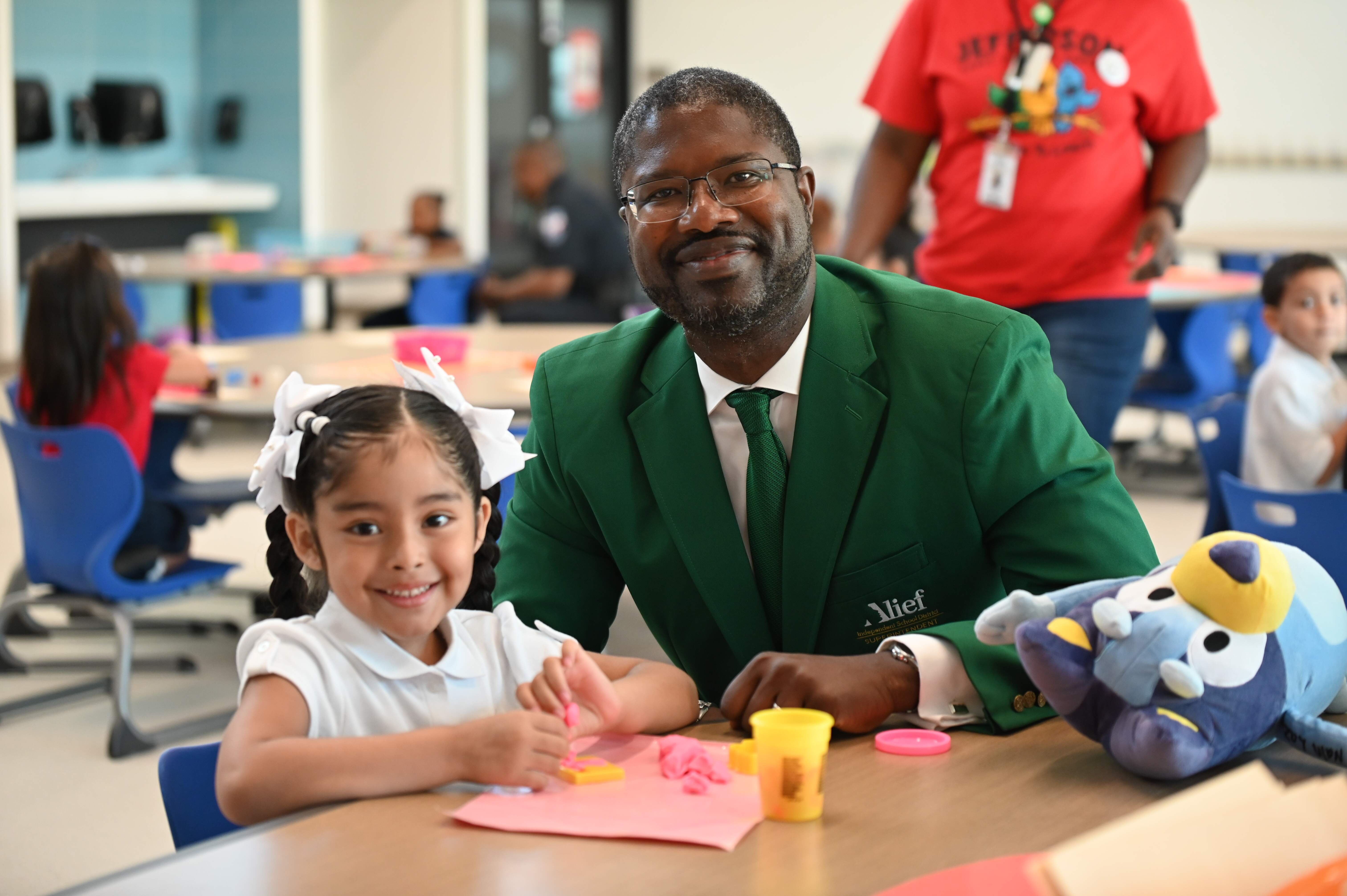 A man in a green suit sits with a child at a table, both smiling. Nearby, another child sits in a chair.