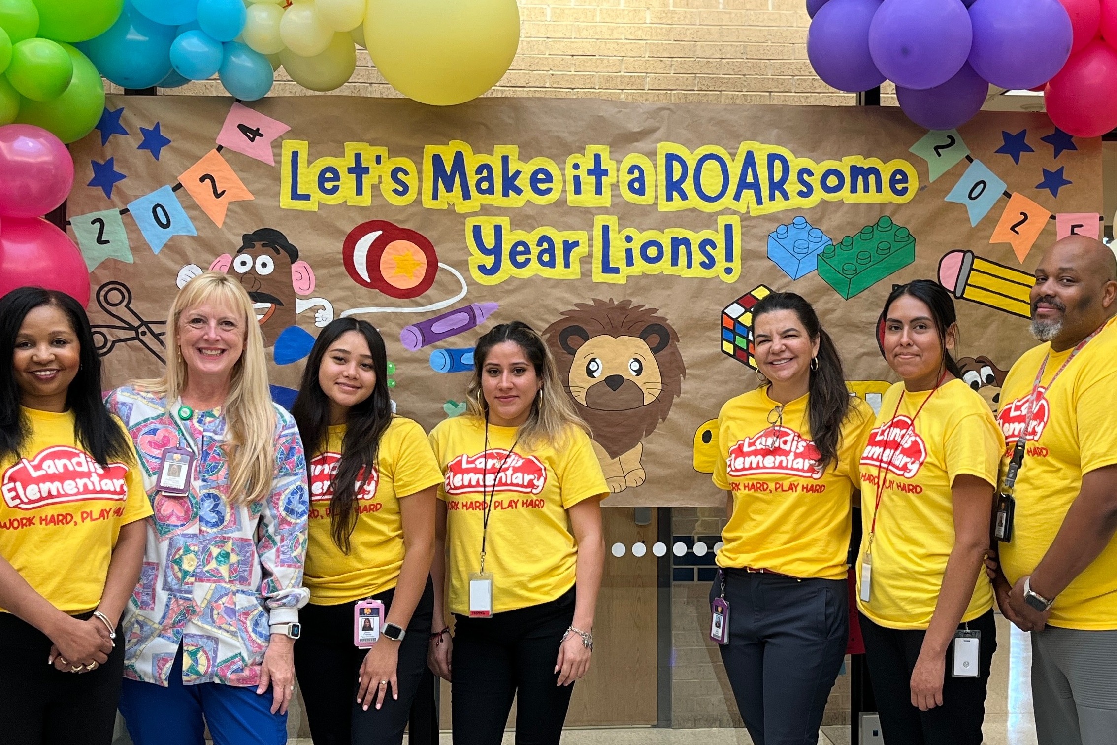 Seven adults in yellow shirts and pants pose in front of a colorful banner with balloons.