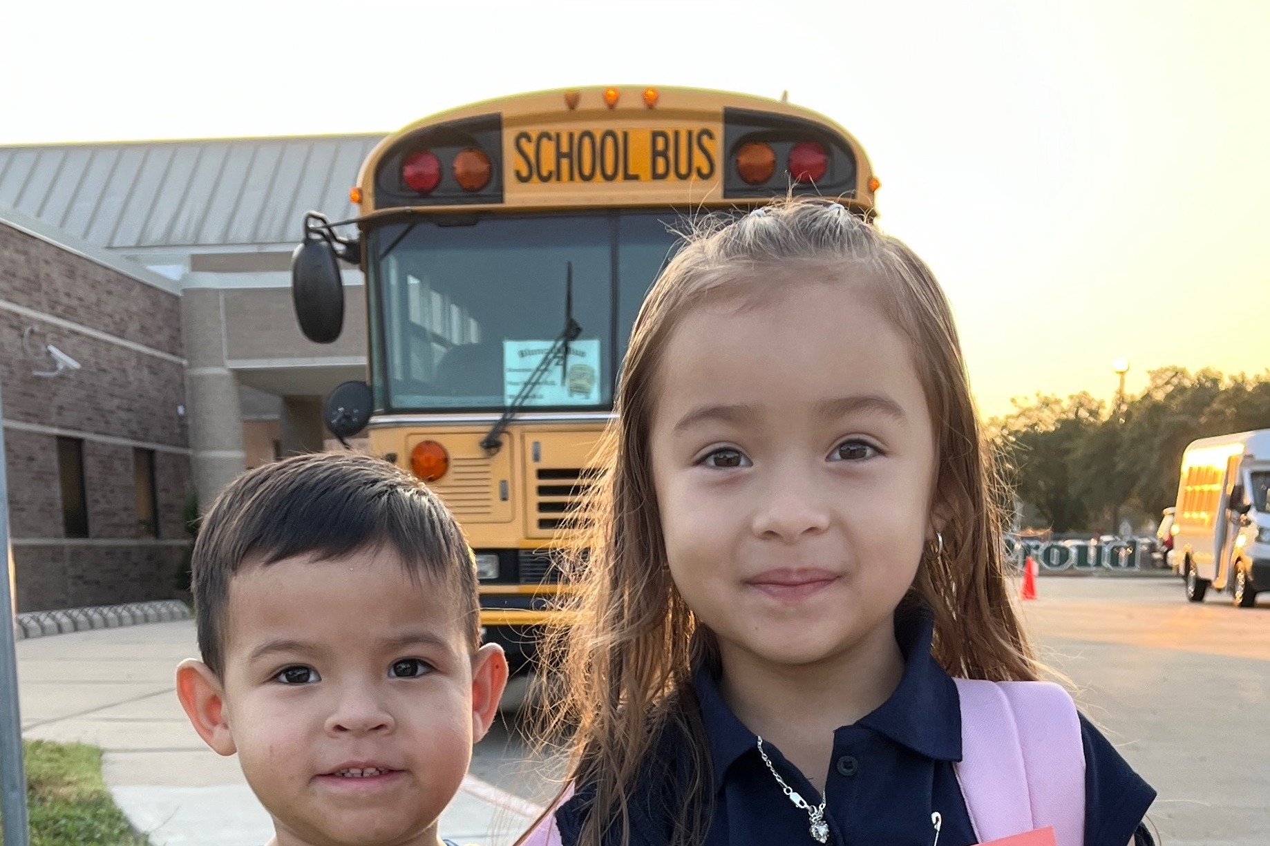 Two young children, a boy and a girl, stand close together in front of a yellow school bus.