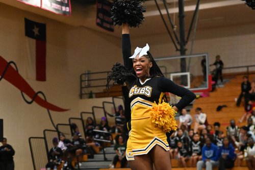 Cheerleader in a black and yellow uniform performing on a stage in a gym. Audience in bleachers.