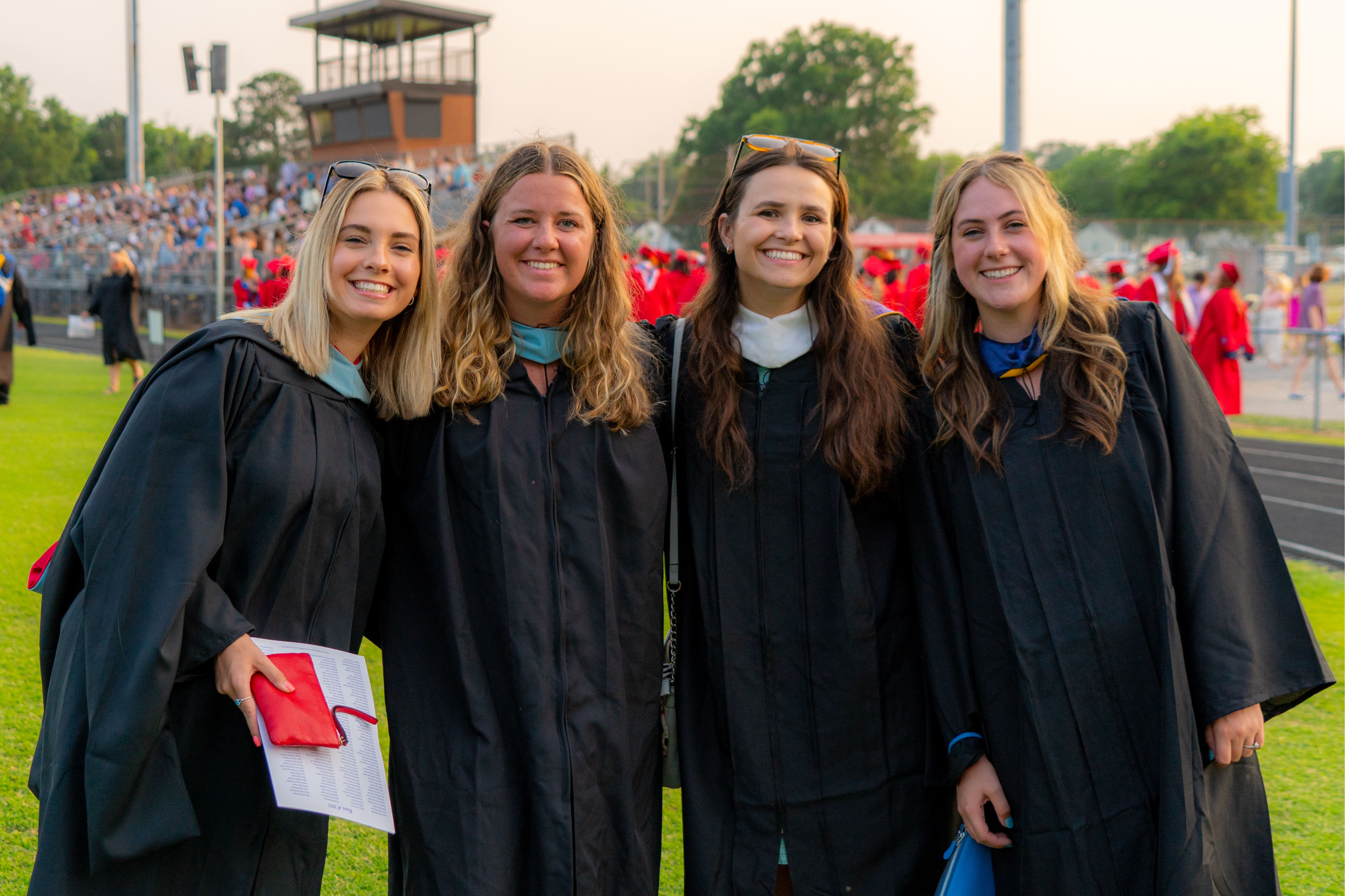 Four white female teachers in their 20s stand together smiling at graduation. They are wearing their black college graduation gowns.