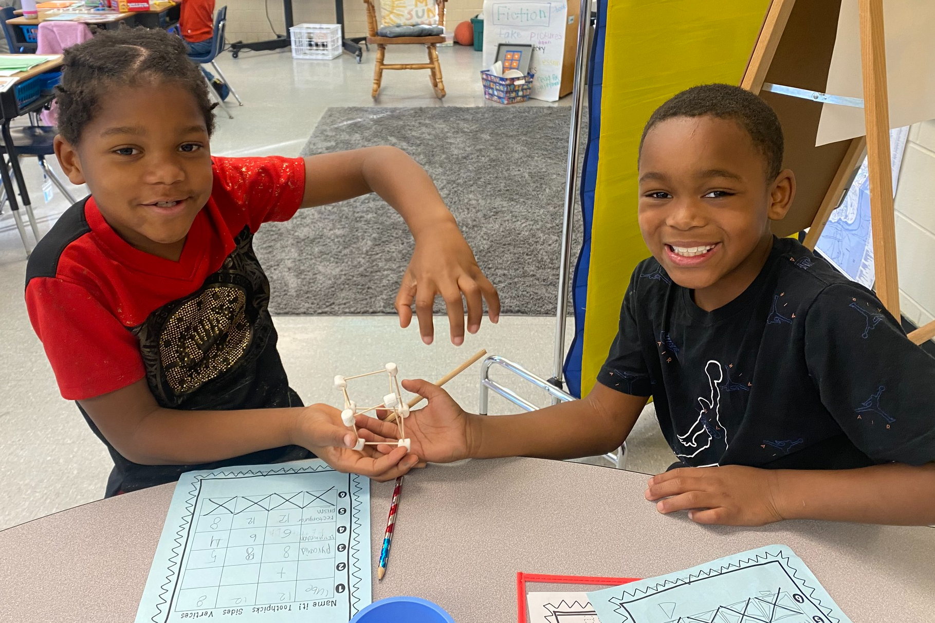 Two elementary-aged black male students proudly hold up a 3D shape they constructed out of marshmallows and toothpicks.