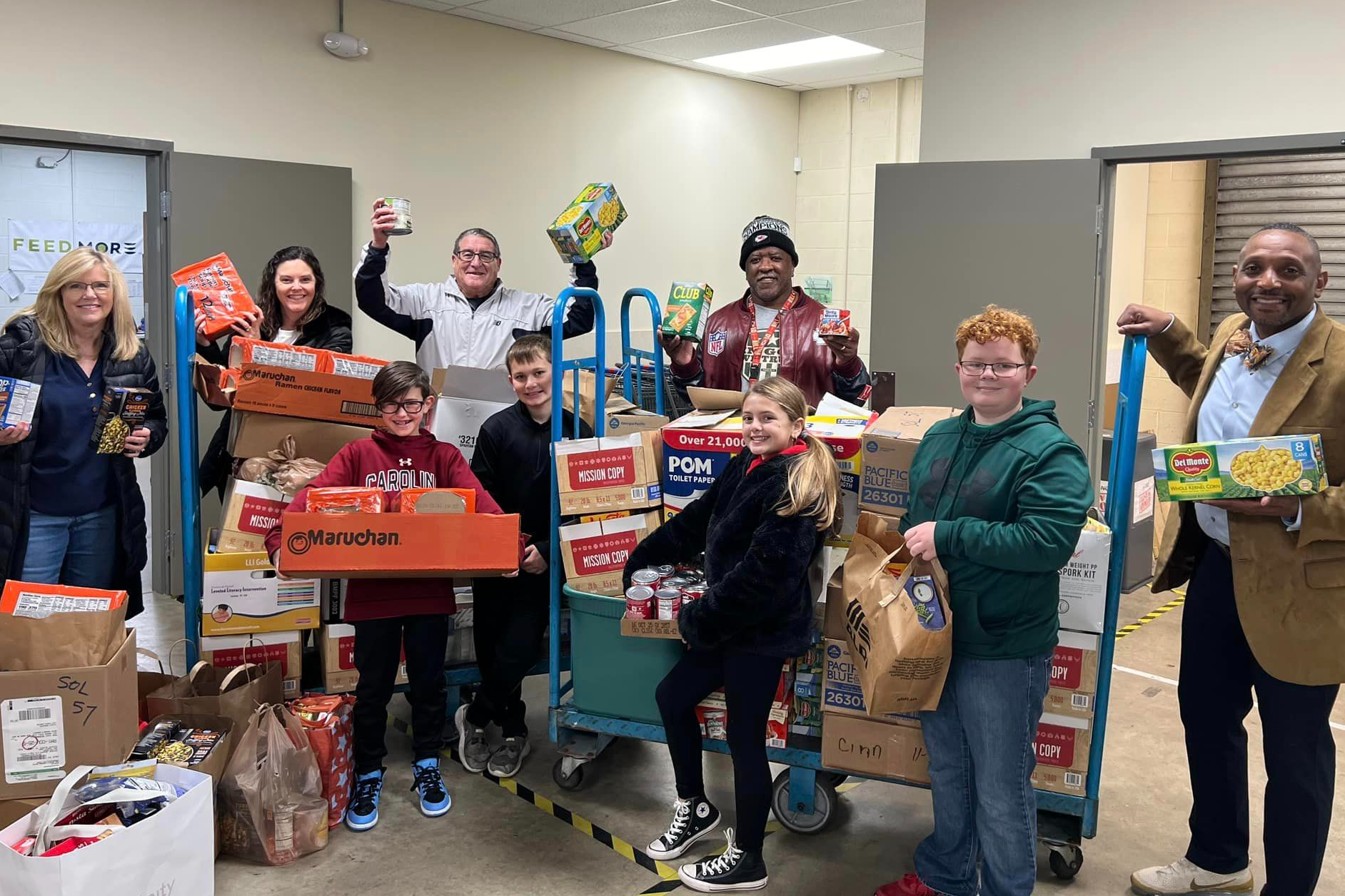 A group of 4 elementary students stand with various adults from the food bank and Tussing Elementary. Around them are numerous bags of nonperishable food they collected as a school to donate.