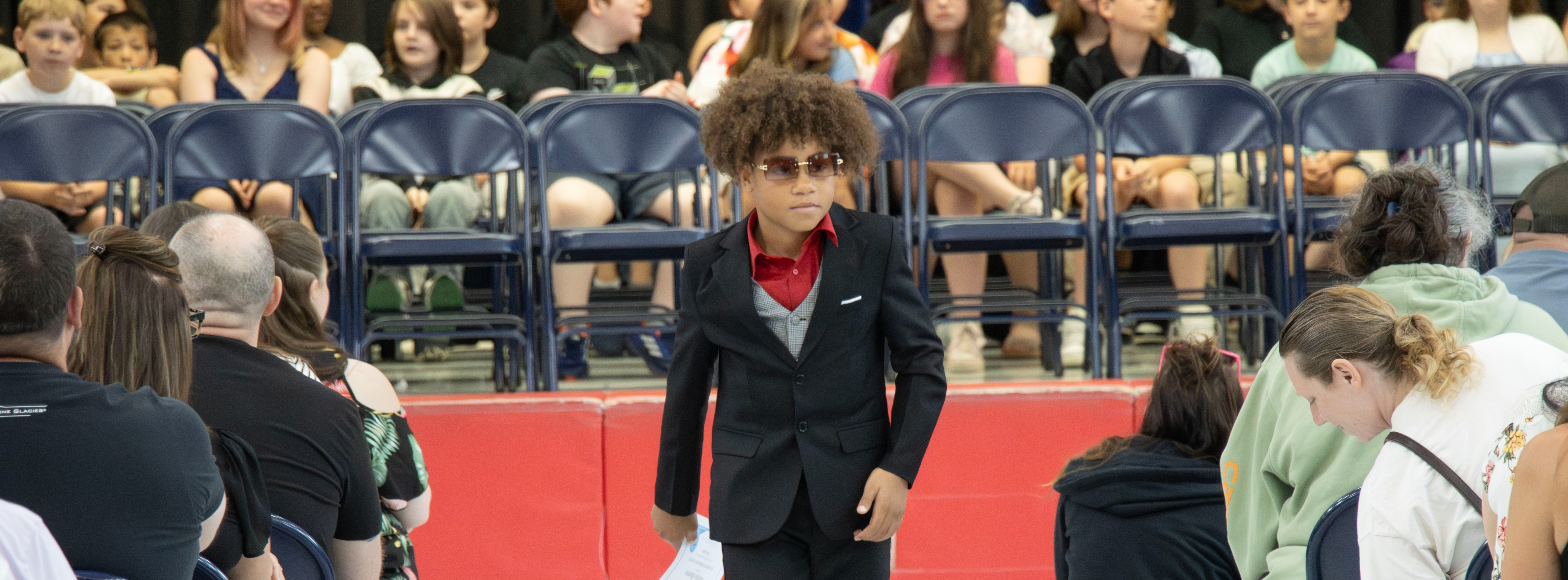 An elementary student wearing a black suit, red shirt and sunglasses walks down the center aisle during a school event as families and other students watch from their seats.An elementary student wearing a black suit, red shirt and sunglasses walks down the center aisle during a school event as families and other students watch from their seats.