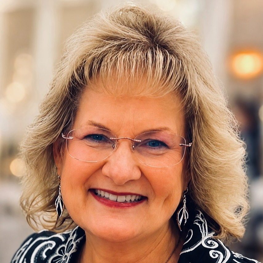 Woman with short blonde hair and glasses smiles, wearing a black and white patterned top.
