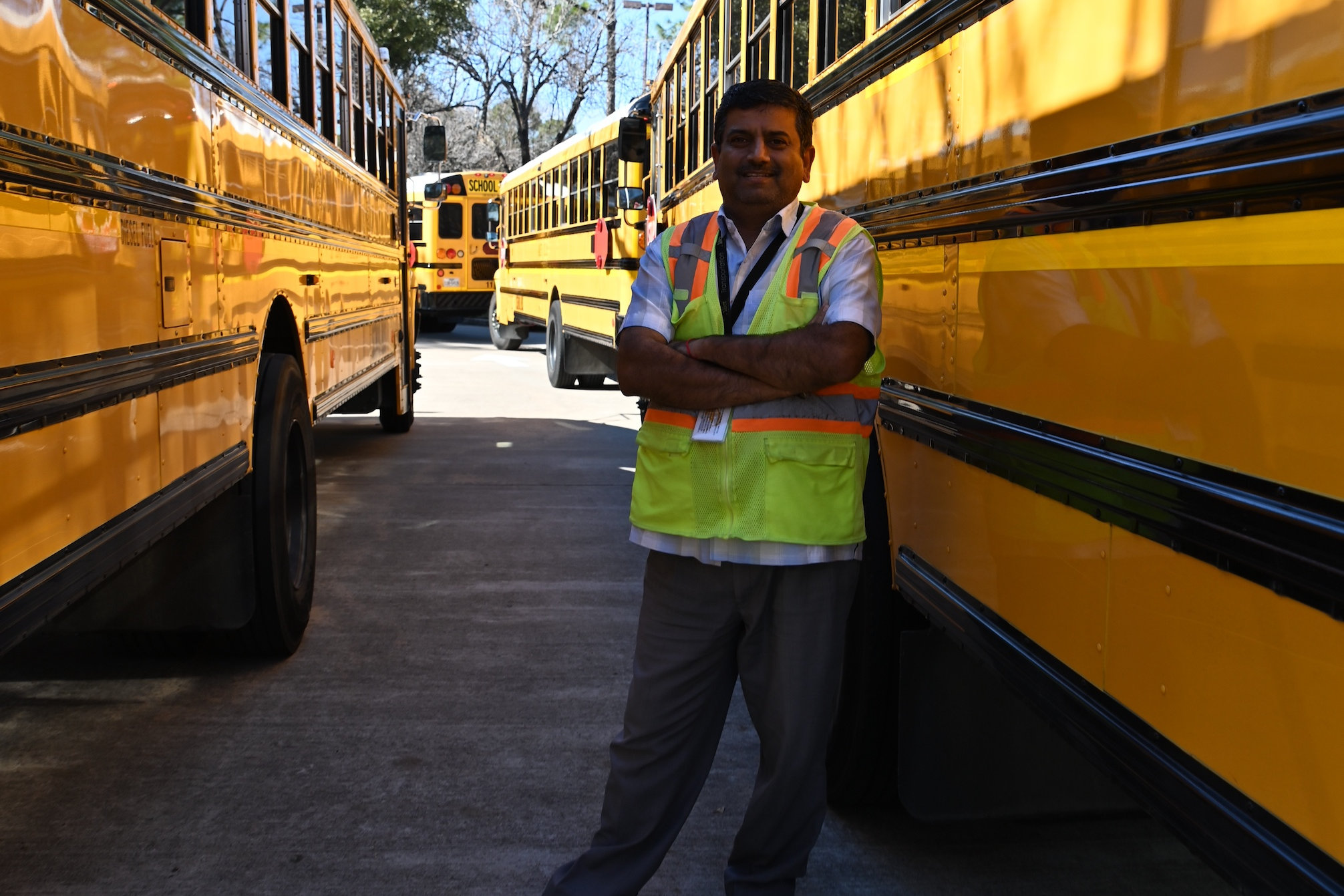 Man in high-visibility vest stands between yellow school buses in a parking lot with trees in the background.