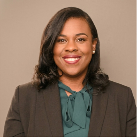 A smiling adult woman with dark hair, wearing a teal blouse and brown blazer, against a gray background.