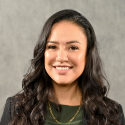 A smiling woman with dark, wavy hair, wearing a green top and a gold necklace, stands against a gray background.
