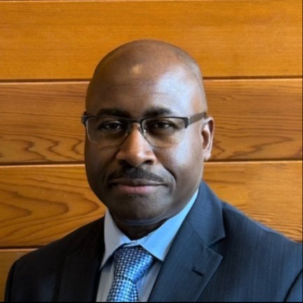 A man wearing a suit, blue tie, and glasses looks at the camera against a wooden background.