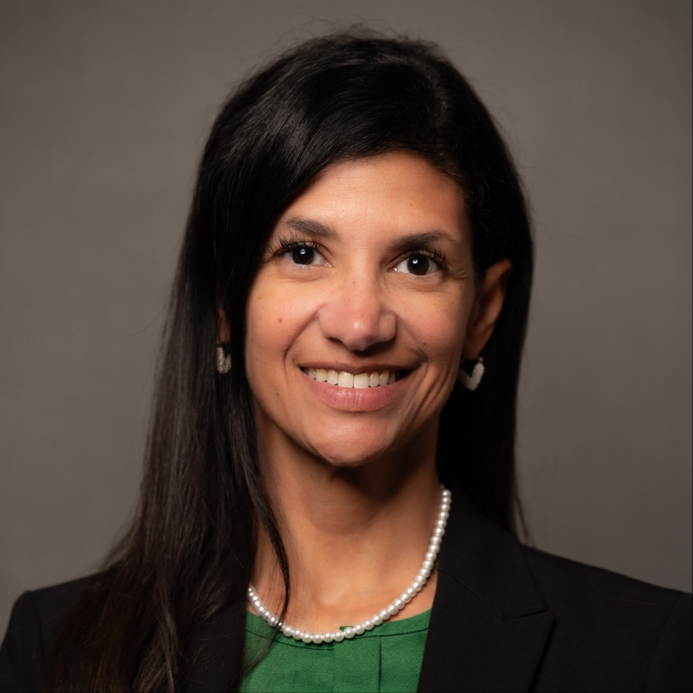 Woman with long dark hair, smiling at camera. Wearing green shirt, pearl necklace, and black blazer.