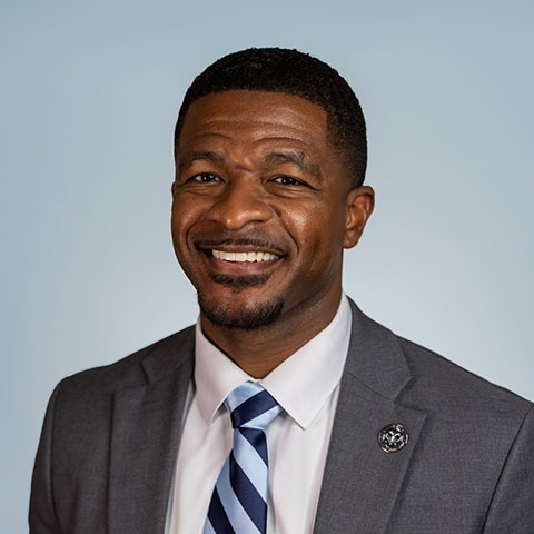 A man in a gray suit with a blue and white striped tie smiles, looking at the camera.