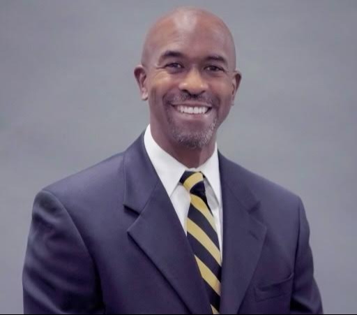 A man in a navy blue suit with a white shirt and a black and yellow striped tie stands with a smile.
