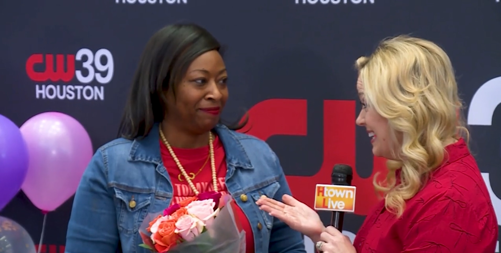 Two women, one in denim and the other in red, converse while holding a bouquet. A microphone and balloons are in the background.