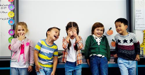 Five small children smiling together in front of a classroom whiteboard