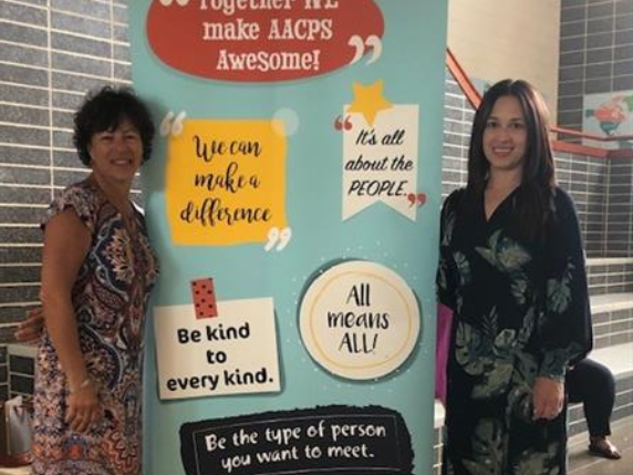 Two women standing in front of a poster with motivational messages and illustrations.