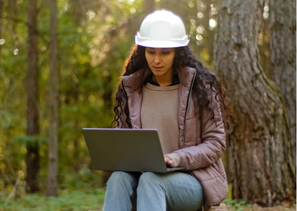 Woman using laptop in woods with hardhat on