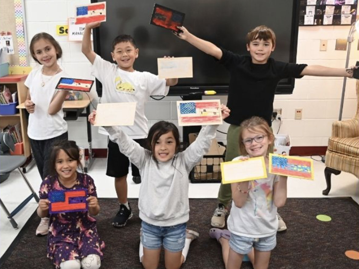 Children in a classroom pose with small flags in their hands. A large TV is visible in the background.