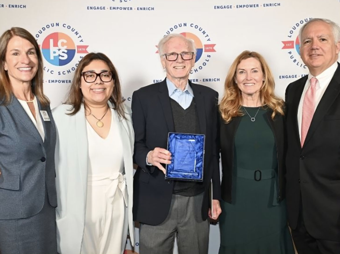 Five people pose for a photo. A man in the center holds a blue award. They are in front of a banner.