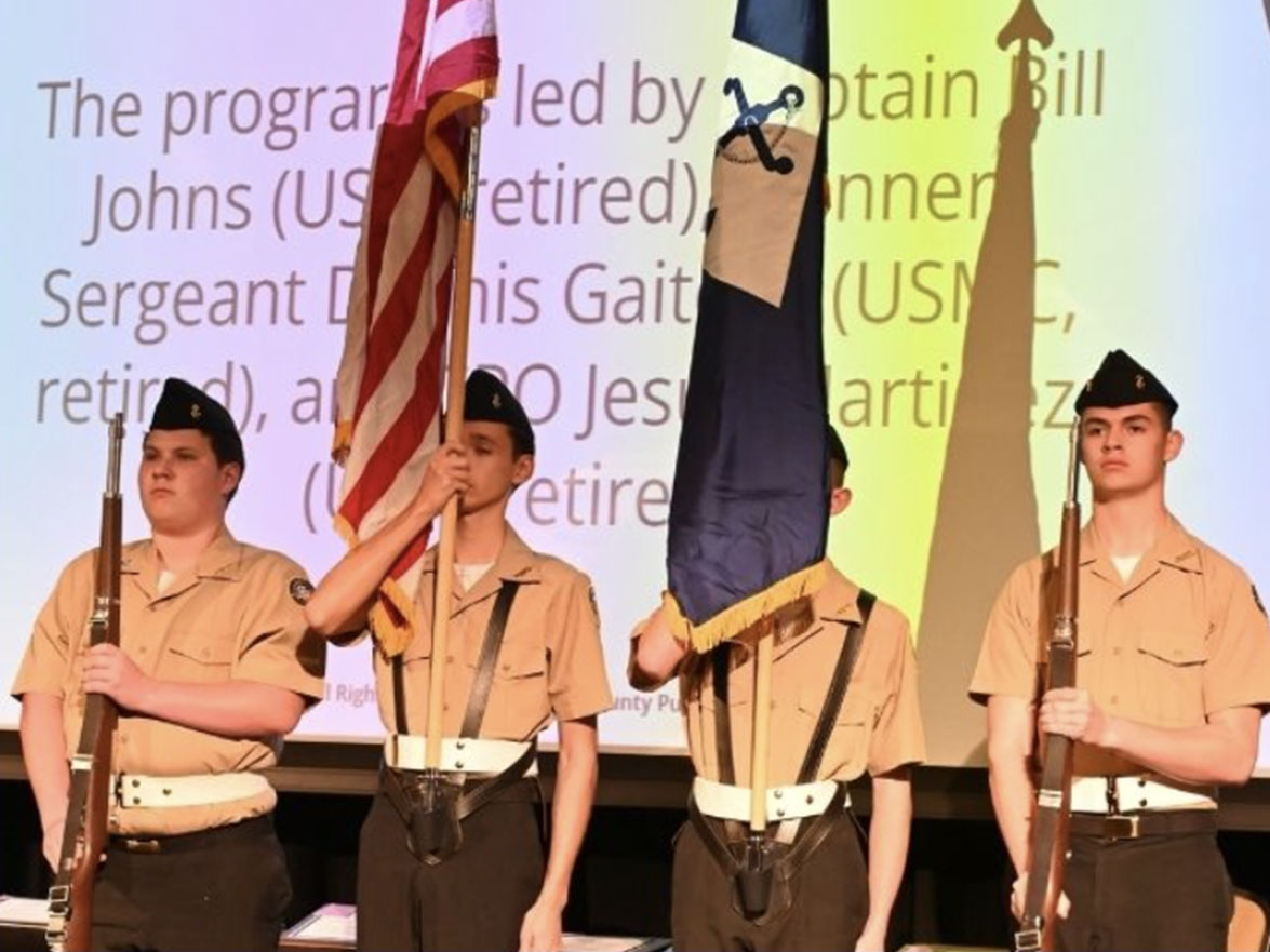Four people in uniform on stage, holding flags. One holds a flagpole while another has a flag.