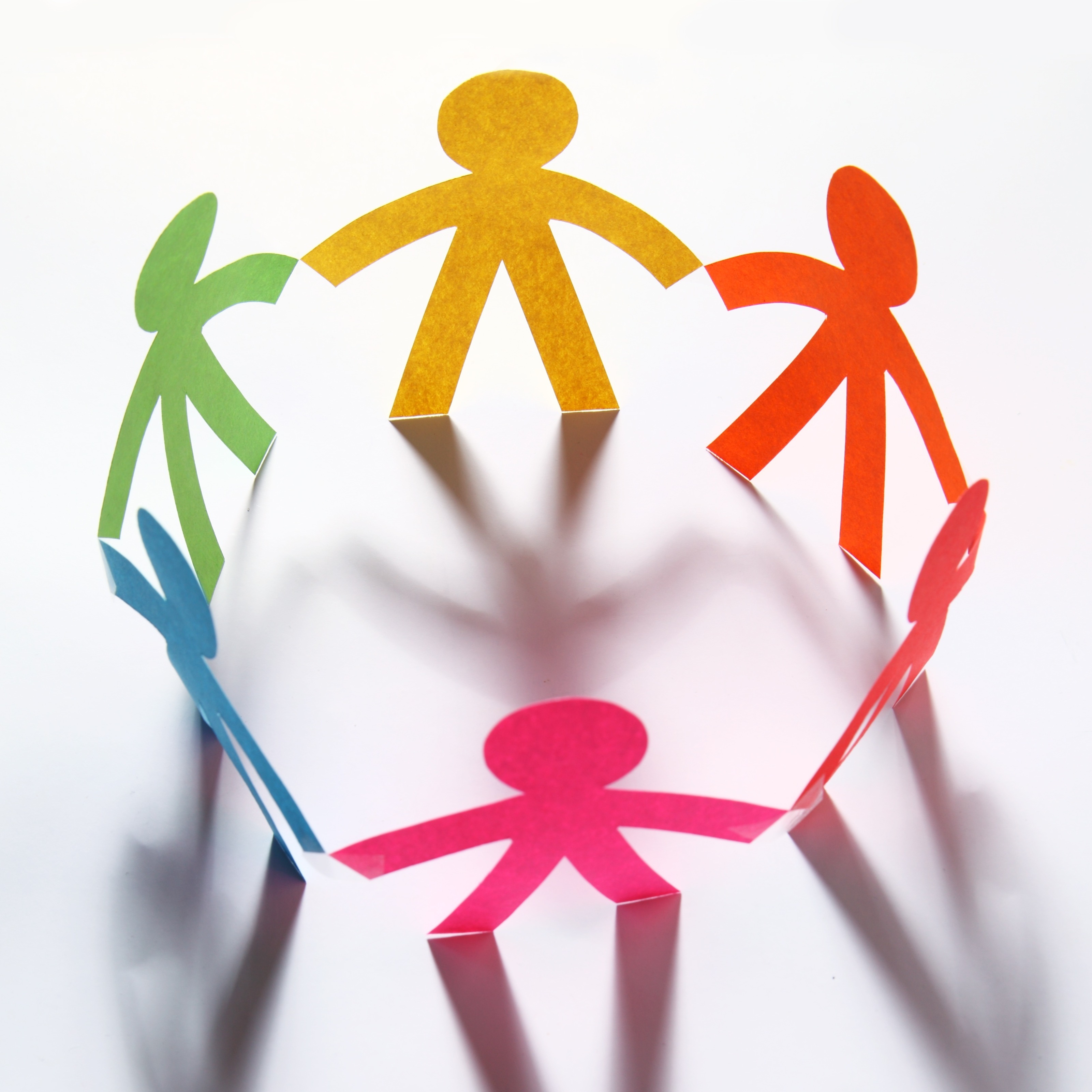 Cut-out paper figures in colors yellow, orange, green, pink, and blue form a circle. Shadows fall on white surface.
