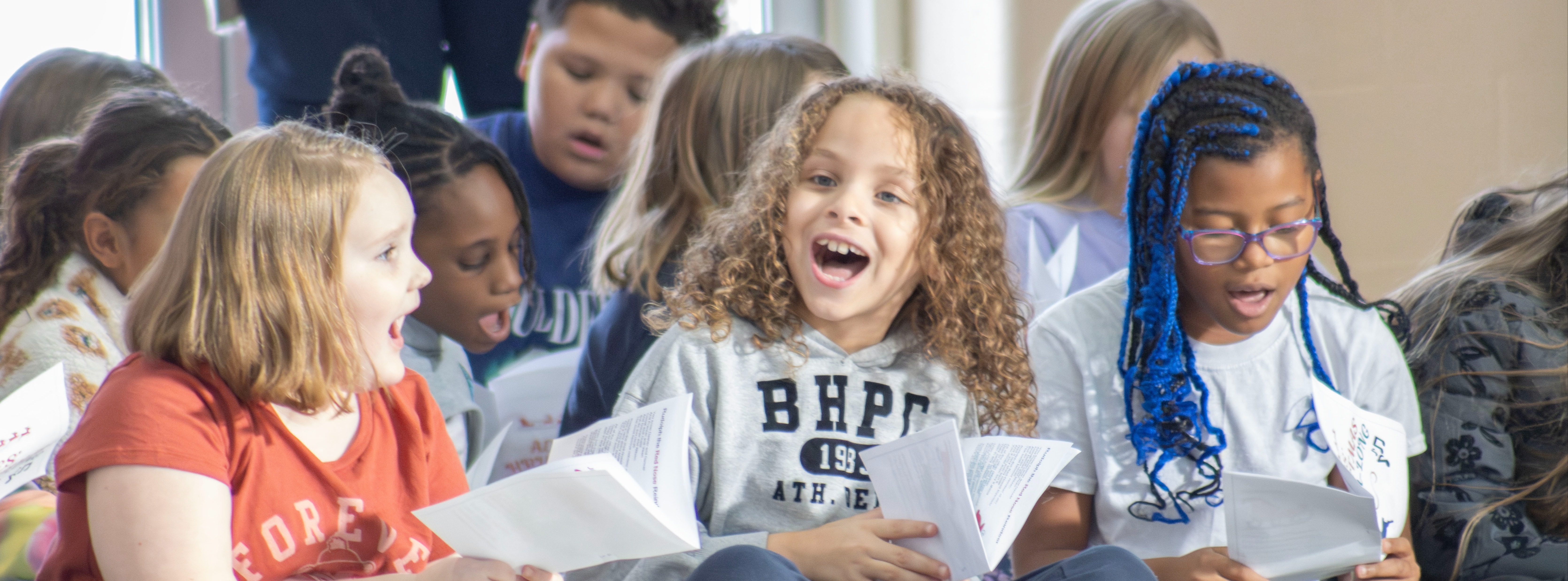 A group of elementary students sit together on the floor holding papers and singing. Three students in the foreground sing enthusiastically while other students participate behind them.