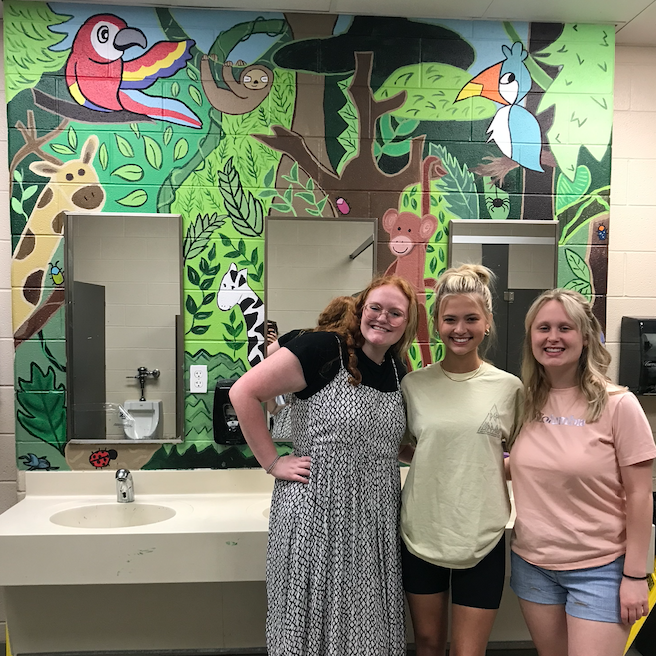 female students and teacher on the boys' restroom sinks displaying the wall painted in a colorful jungle theme