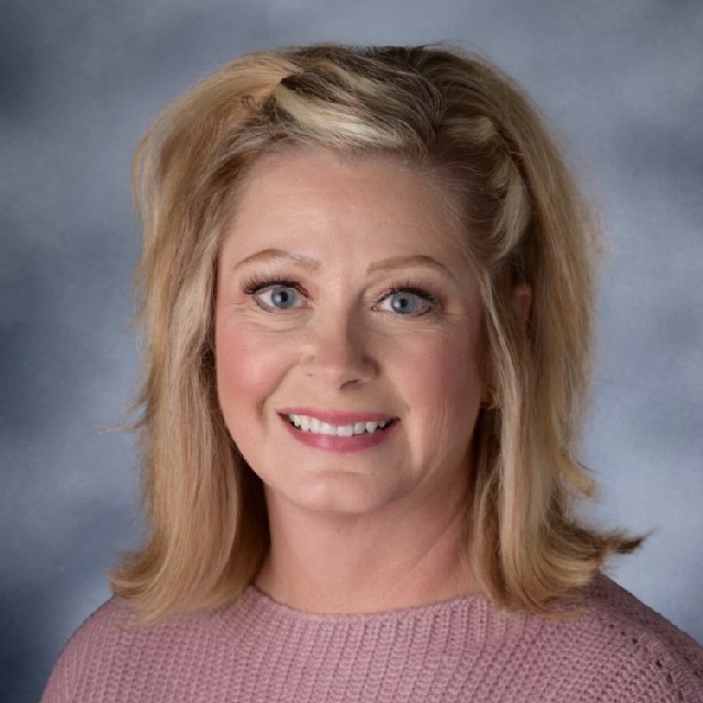 A woman with blonde hair and blue eyes smiles at the camera. She wears a light pink sweater.