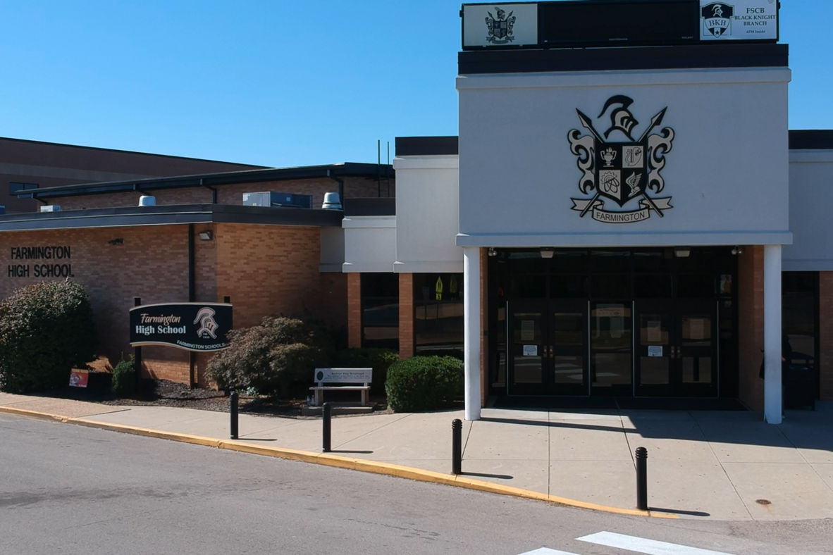 Building with emblem, double doors, and bench outside. Buildings in background. Sidewalk, shrubs, and bollards.