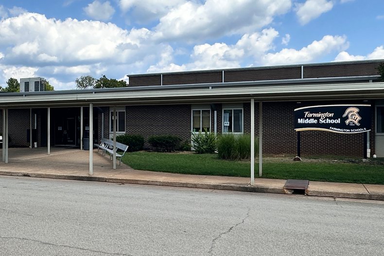 An empty street in front of a school with an open porch. A blue sky with clouds and a flagpole.