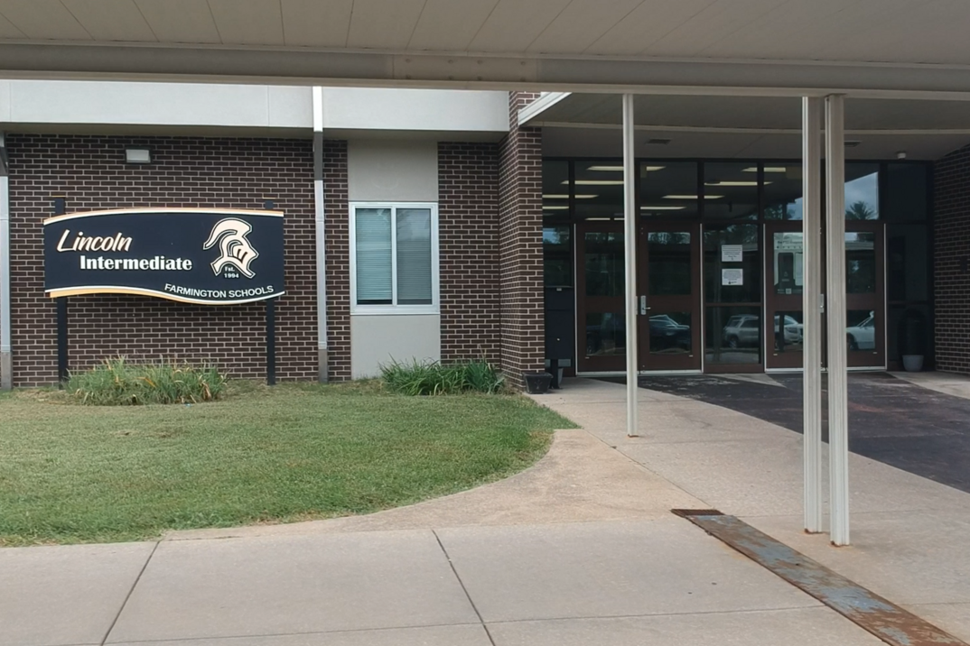 A school entrance with a black sign above the entrance, brick walls, windows, and a grassy area.