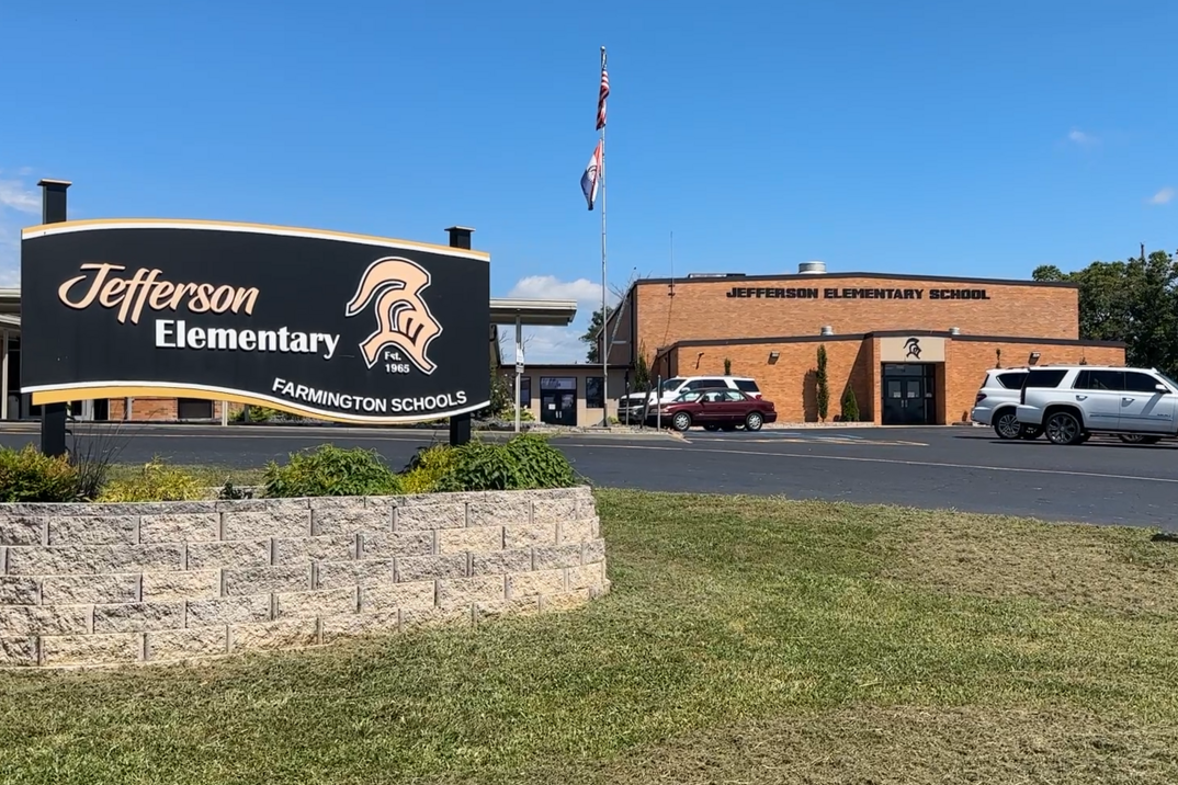 Entrance to Jefferson Elementary. Sign with logo. Cars parked in front. Building with flag. Trees in the background.