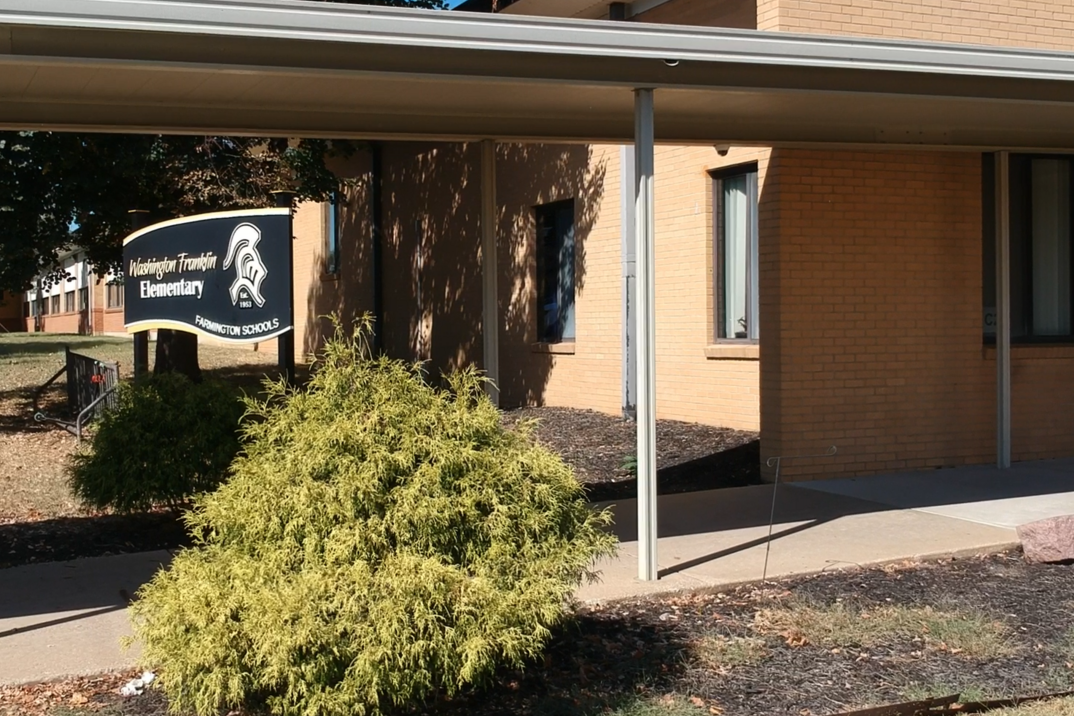 A building exterior features a canopy, sign with a logo, and a bush. A sidewalk leads to the entrance.